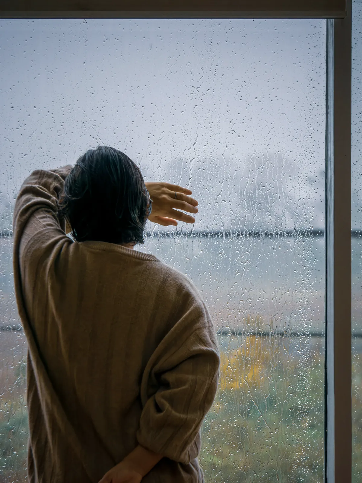Une personne de dos regarde la pluie derrière une grande fenêtre